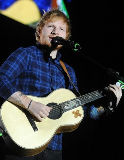 Ed Sheeran singing and playing an acoustic guitar on stage with colorful lights behind him.