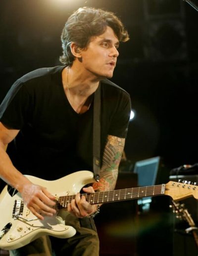 John Mayer playing an electric guitar on stage, wearing a black shirt with a tattooed arm.