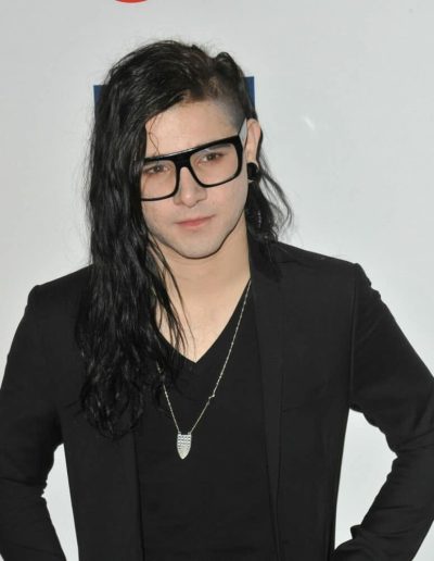 Skrillex wearing glasses and a black suit, standing in front of a plain background.