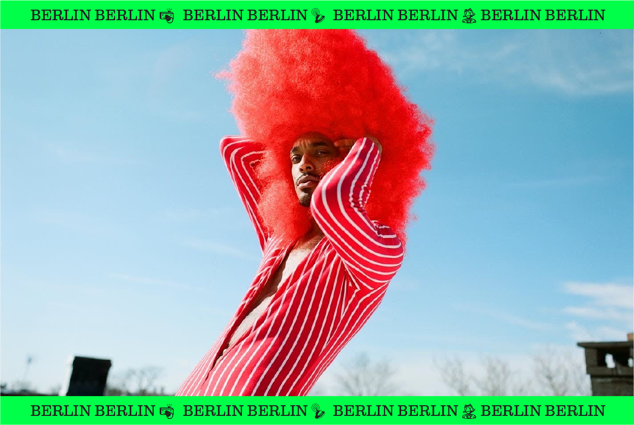 Image of Berlin Berlin in a huge red frizzy wig and wearing a red and white striped top.